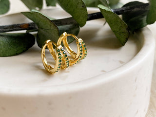 Gold hoop earrings with green gemstones on a marble surface with green leaves in the background