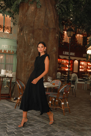 Woman in a black dress walking in an outdoor setting with tables and chairs.