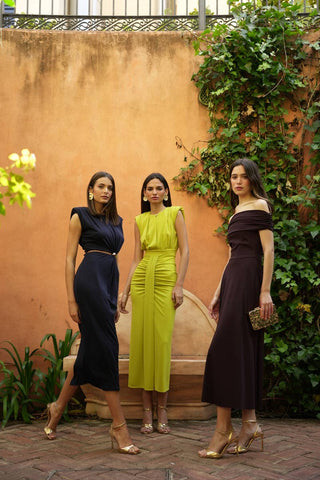Three women in stylish outfits standing against a textured wall with plants.