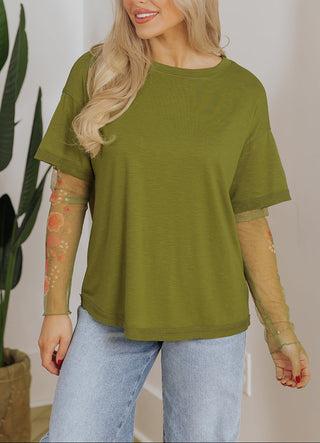 Woman wearing a green t-shirt and blue jeans standing indoors with a plant in the background